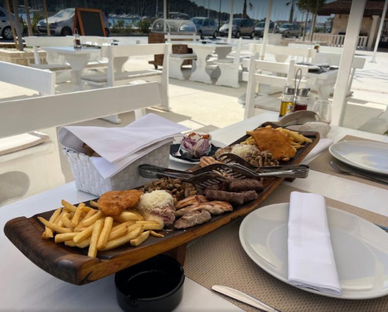 plate potatoes on a wooden platter breaded meat čevapi burgers grated cheese mushrooms white table plates sea view restaurant rogoznica
